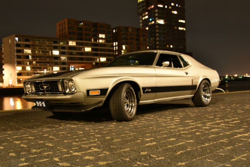 Sfeervolle avondfoto van de te huren Ford Mustang muscle car op de kade met uitzicht op de skyline van Rotterdam.
